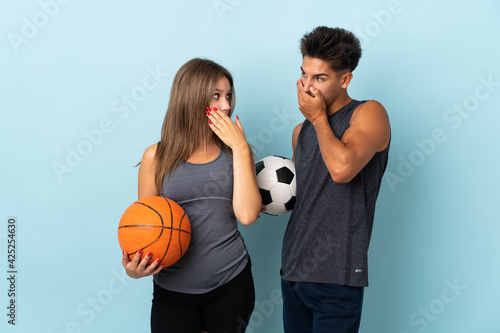 Young couple playing football and basketball isolated on blue background covering mouth with hands for saying something inappropriate