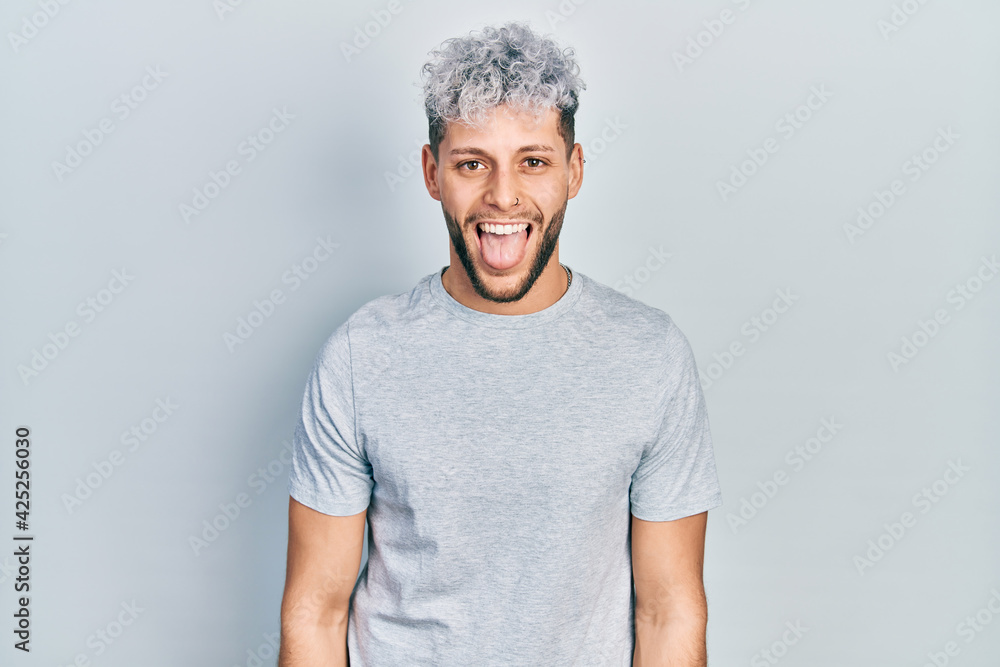 Young hispanic man with modern dyed hair wearing casual grey t shirt sticking tongue out happy with funny expression. emotion concept. Young hispanic man with modern dyed hair wearing casual grey t shirt sticking tongue out happy with funny expression. emotion concept.