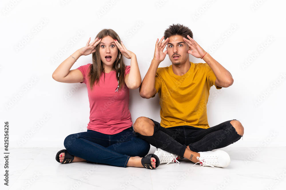 Young couple sitting on the floor isolated on white background with surprise expression