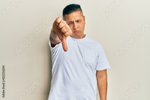 Young latin man wearing casual white t shirt looking unhappy and angry showing rejection and negative with thumbs down gesture. bad expression.