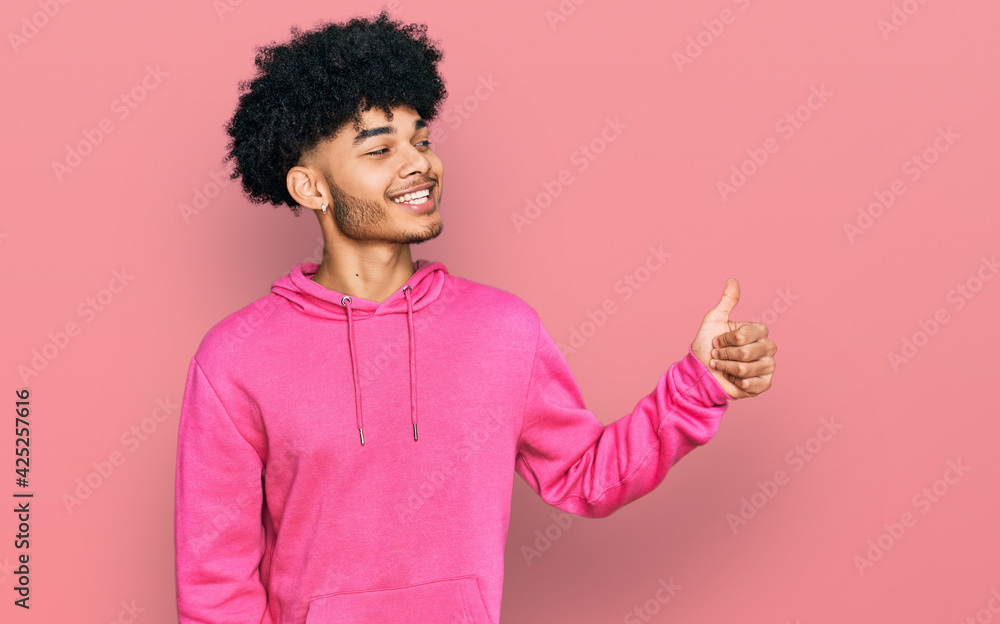 Young african american man with afro hair wearing casual pink sweatshirt looking proud, smiling doing thumbs up gesture to the side Young african american man with afro hair wearing casual pink sweatshirt looking proud, smiling doing thumbs up gesture to the side