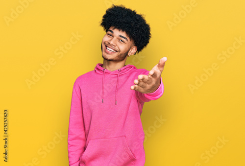 Young african american man with afro hair wearing casual pink sweatshirt smiling cheerful offering palm hand giving assistance and acceptance.