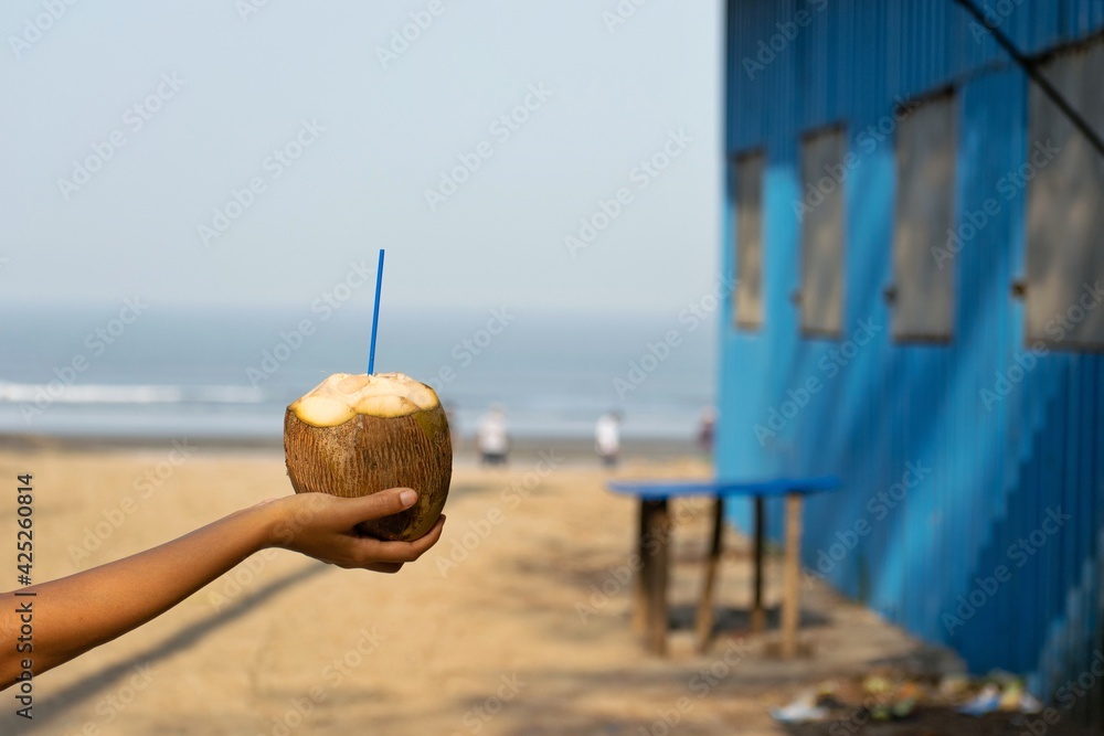 A picture of a hand of a woman holding a coconut with a straw in it at beach in the morning. Drinking fresh coconut water is considered healthy superfood, detox consumed after workout, in summer, hot.