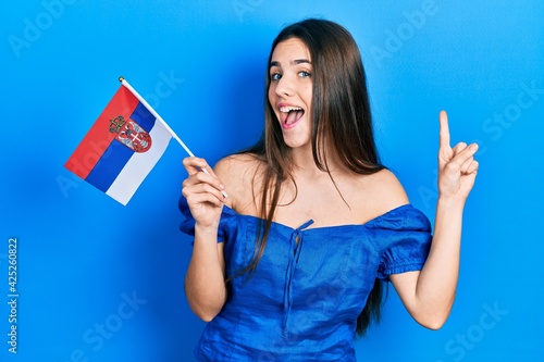 Young brunette teenager holding serbia flag smiling with an idea or question pointing finger with happy face  number one