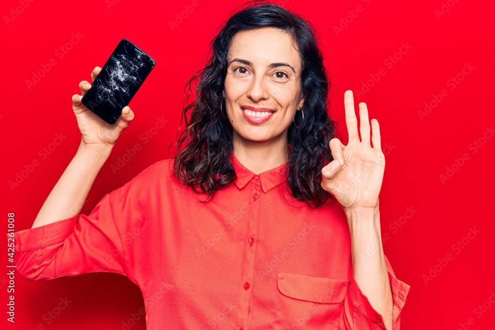 Naklejka premium Young beautiful hispanic woman holding broken smartphone showing cracked screen doing ok sign with fingers, smiling friendly gesturing excellent symbol