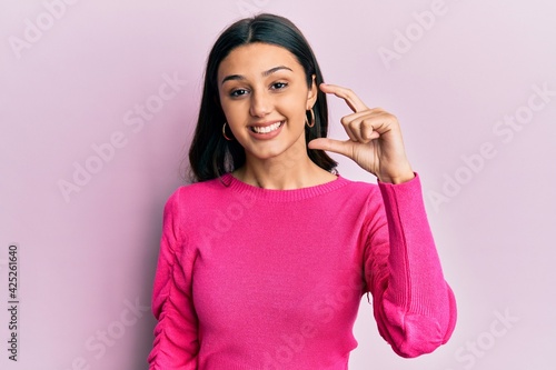 Young hispanic woman wearing casual clothes smiling and confident gesturing with hand doing small size sign with fingers looking and the camera. measure concept.
