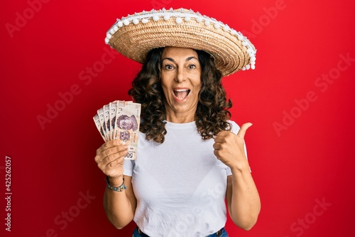 Middle age hispanic woman wearing mexican hat holding pesos banknotes pointing thumb up to the side smiling happy with open mouth © Krakenimages.com