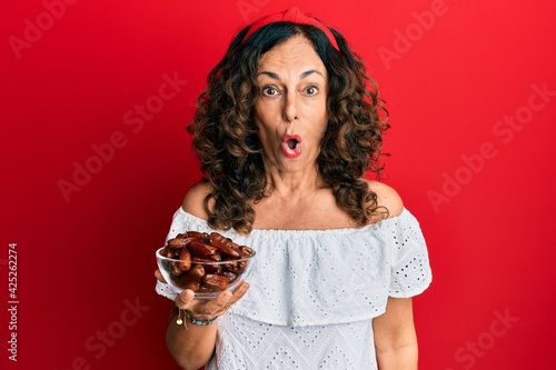 Middle age hispanic woman holding bowl with dates scared and amazed with open mouth for surprise, disbelief face