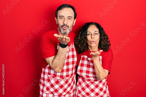 Middle age couple of hispanic woman and man wearing cook apron looking at the camera blowing a kiss with hand on air being lovely and sexy. love expression.