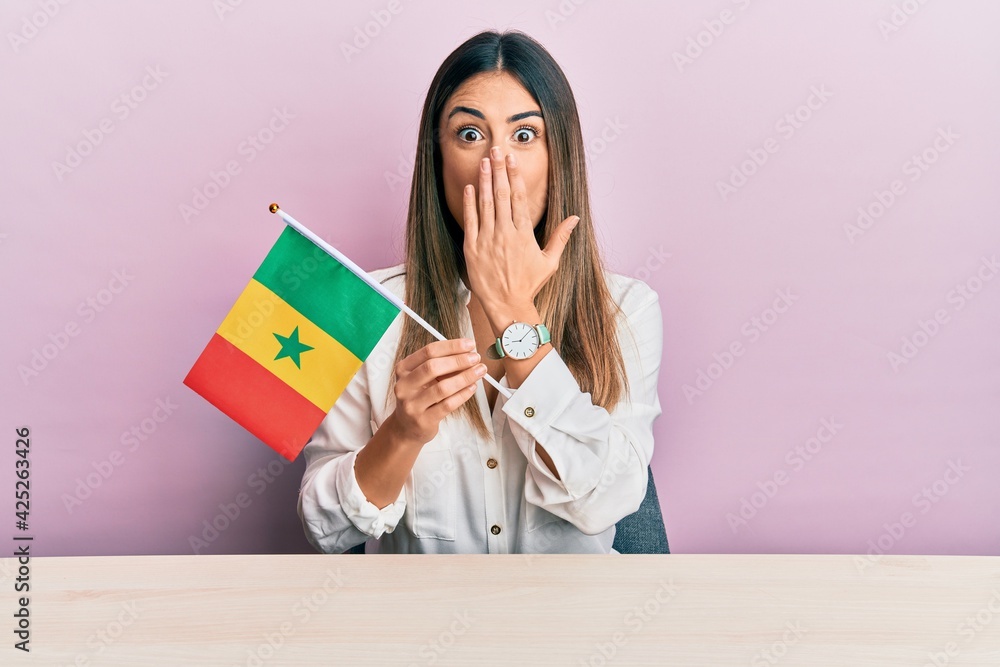 Young hispanic woman holding senegal flag sitting on the table covering mouth with hand, shocked and afraid for mistake. surprised expression Young hispanic woman holding senegal flag sitting on the table covering mouth with hand, shocked and afraid for mistake. surprised expression
