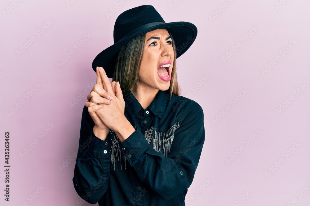 Fototapeta premium Young hispanic woman wearing cowgirl style doing pistol gesture with hands angry and mad screaming frustrated and furious, shouting with anger. rage and aggressive concept.