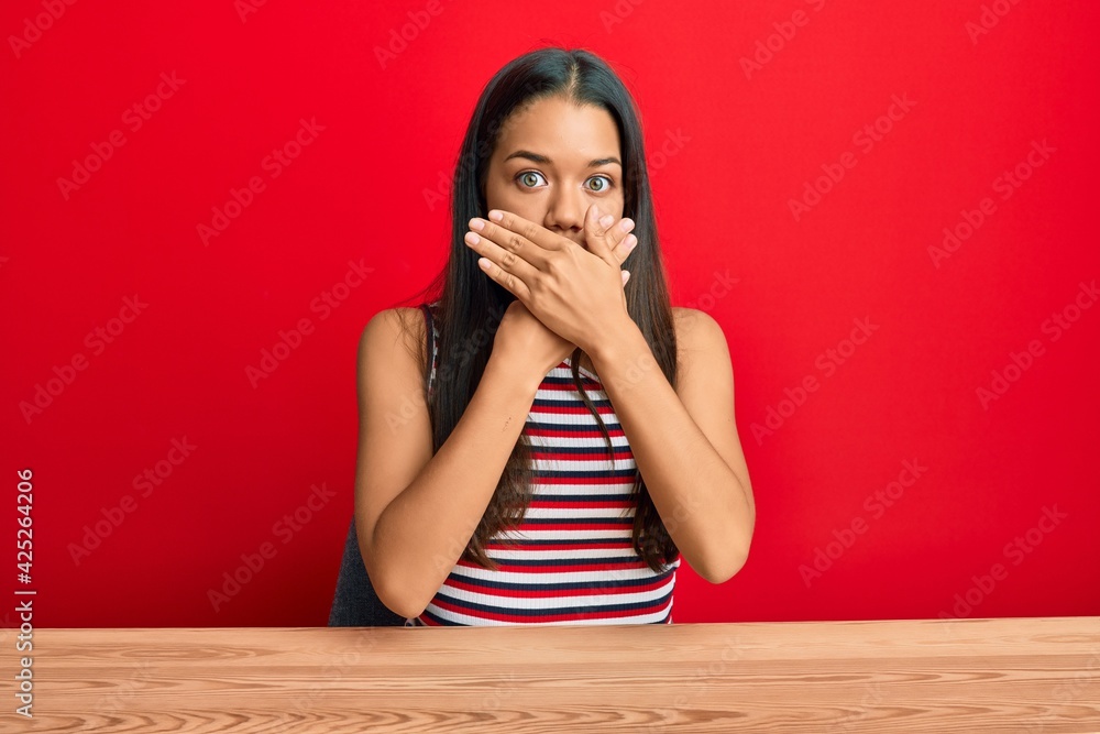 Beautiful hispanic woman wearing casual clothes sitting on the table shocked covering mouth with hands for mistake. secret concept. Beautiful hispanic woman wearing casual clothes sitting on the table shocked covering mouth with hands for mistake. secret concept.