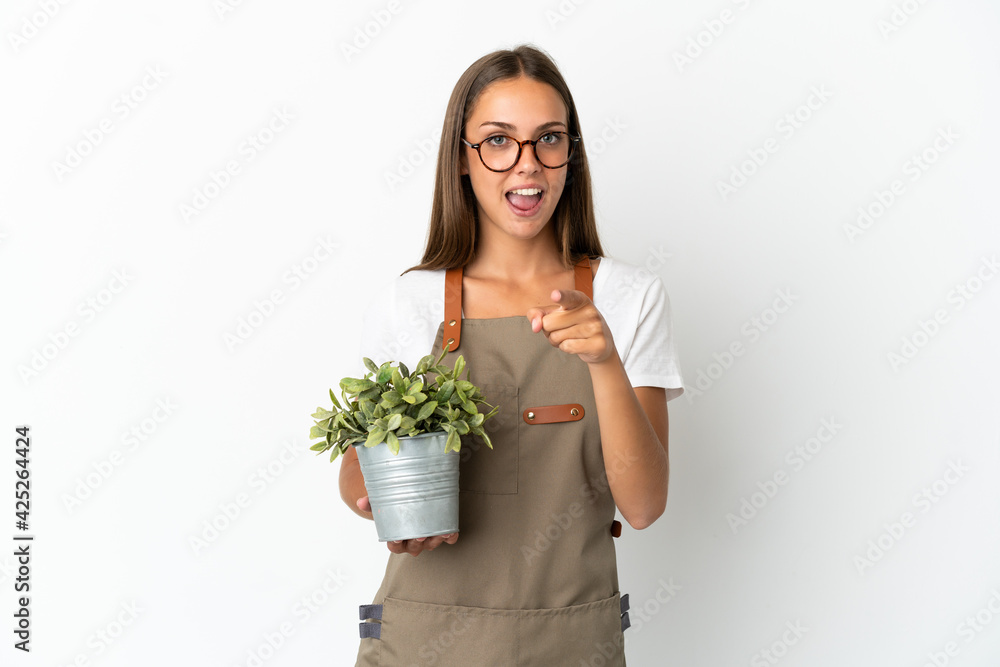 Gardener girl holding a plant over isolated white background surprised and pointing front Gardener girl holding a plant over isolated white background surprised and pointing front