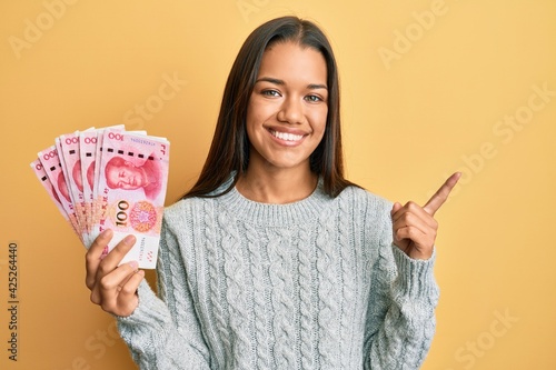 Beautiful hispanic woman holding 100 yuan chinese banknotes smiling happy pointing with hand and finger to the side
