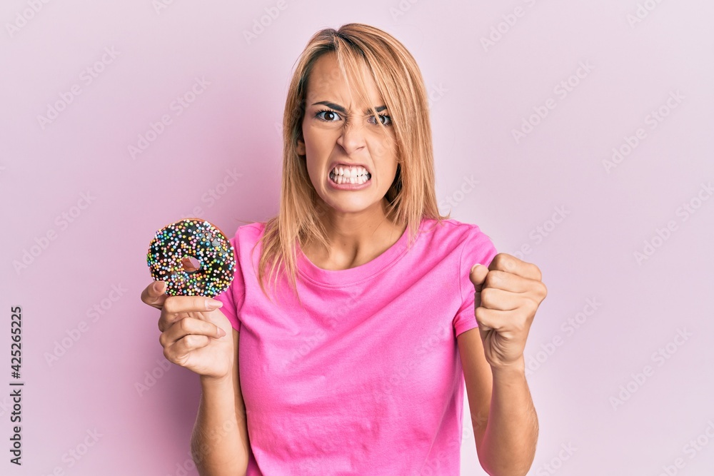 Beautiful blonde woman holding donut annoyed and frustrated shouting with anger, yelling crazy with anger and hand raised Beautiful blonde woman holding donut annoyed and frustrated shouting with anger, yelling crazy with anger and hand raised