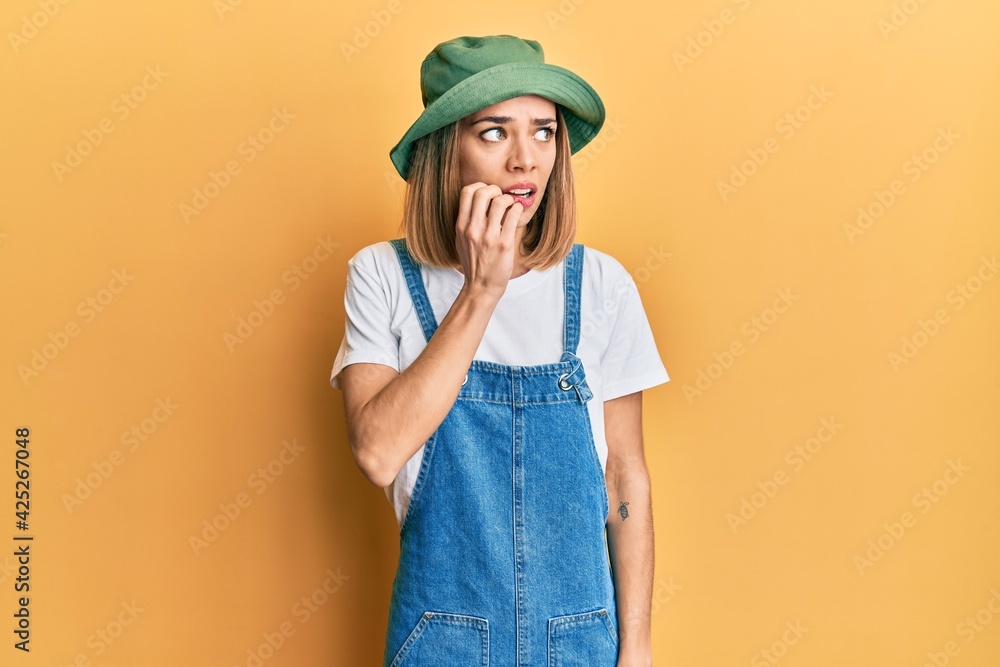 Young caucasian blonde woman wearing denim jumpsuit and hat with 90s style looking stressed and nervous with hands on mouth biting nails. anxiety problem. Young caucasian blonde woman wearing denim jumpsuit and hat with 90s style looking stressed and nervous with hands on mouth biting nails. anxiety problem.