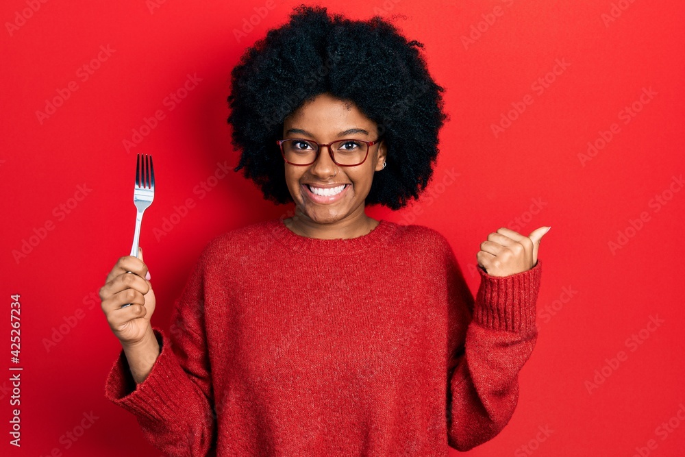 Young african american woman holding one silver fork pointing thumb up to the side smiling happy with open mouth Young african american woman holding one silver fork pointing thumb up to the side smiling happy with open mouth