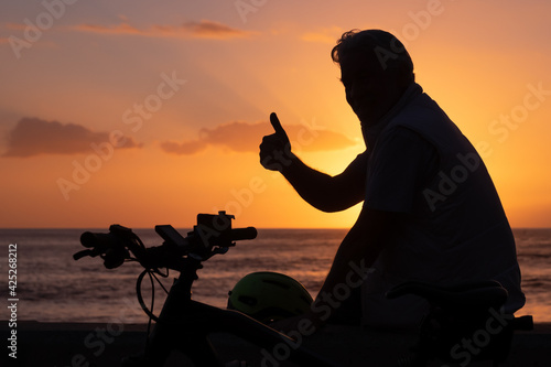 Silhouette of mature man sitting near his bicycle at the beach with a magnificent orange sunset. Thumb up looking at the camera