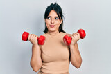 Young hispanic woman wearing sportswear using dumbbells smiling looking to the side and staring away thinking.