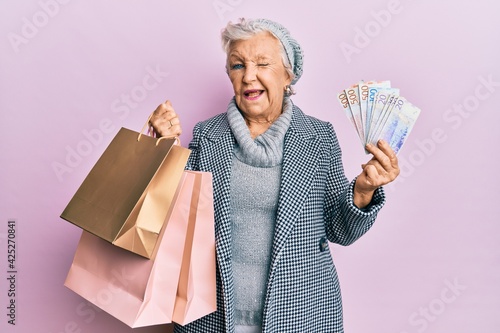 Senior grey-haired woman holding shopping bags and swedish krona banknotes winking looking at the camera with sexy expression, cheerful and happy face.