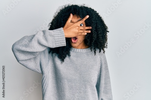 African american woman with afro hair wearing casual winter sweater peeking in shock covering face and eyes with hand, looking through fingers afraid © Krakenimages.com