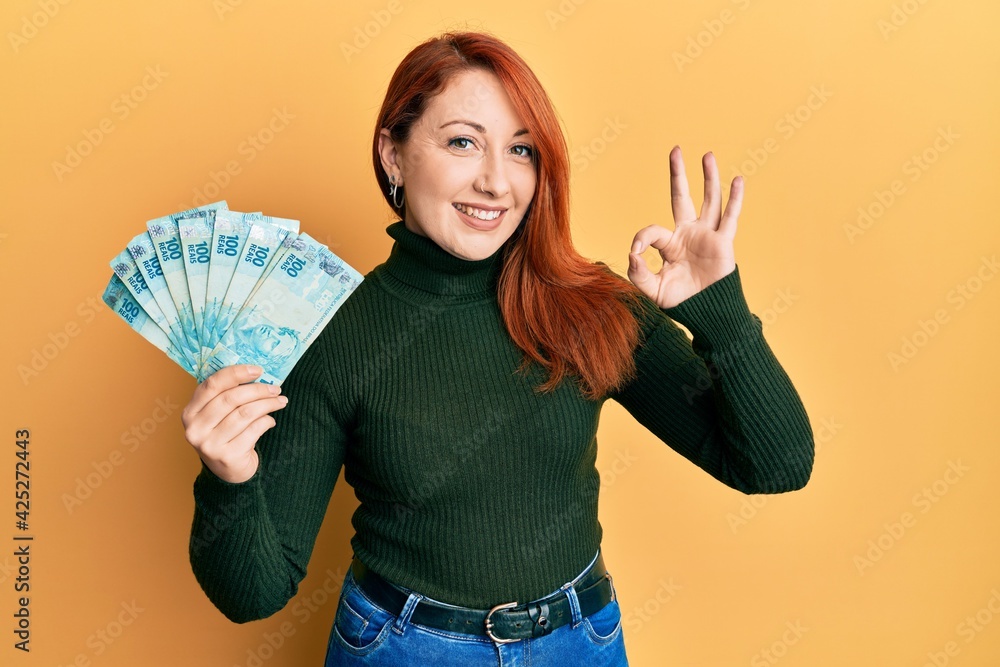 Beautiful redhead woman holding 100 brazilian real banknotes doing ok sign with fingers, smiling friendly gesturing excellent symbol Beautiful redhead woman holding 100 brazilian real banknotes doing ok sign with fingers, smiling friendly gesturing excellent symbol