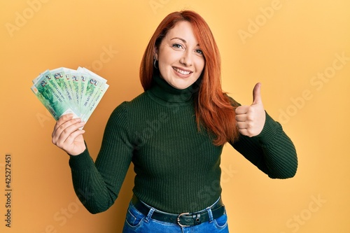 Beautiful redhead woman holding 5 singapore dollars banknotes smiling happy and positive  thumb up doing excellent and approval sign