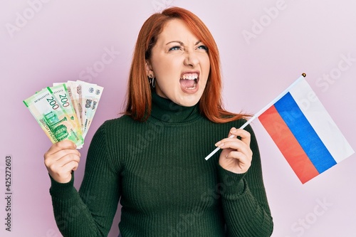 Beautiful redhead woman holding russian ruble banknotes and russia flag angry and mad screaming frustrated and furious, shouting with anger. rage and aggressive concept.