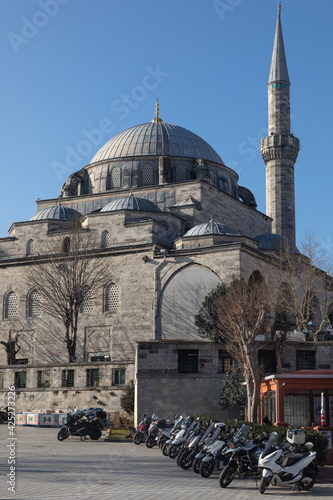 A vertical photo of a mosque made of white stone with high minarets. At the very top there are large dome, below there are small domes. Below there is a parking for motorbikes on white paving stones. photo