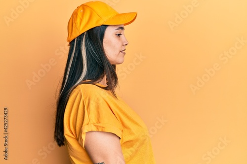 Young hispanic woman wearing delivery uniform and cap looking to side, relax profile pose with natural face with confident smile.