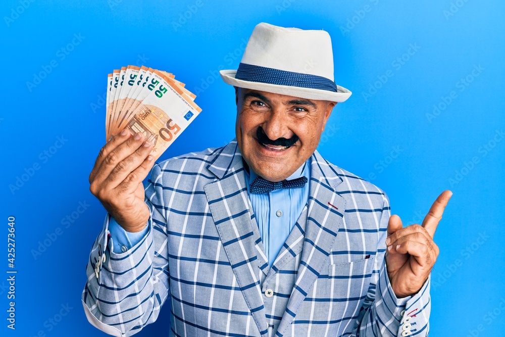 Mature middle east man with mustache wearing elegant vintage style holding euros banknotes smiling happy pointing with hand and finger to the side Mature middle east man with mustache wearing elegant vintage style holding euros banknotes smiling happy pointing with hand and finger to the side