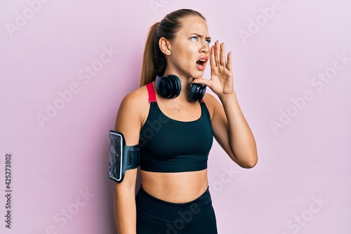 Beautiful young blonde woman wearing gym clothes and using headphones shouting and screaming loud to side with hand on mouth. communication concept. © Krakenimages.com