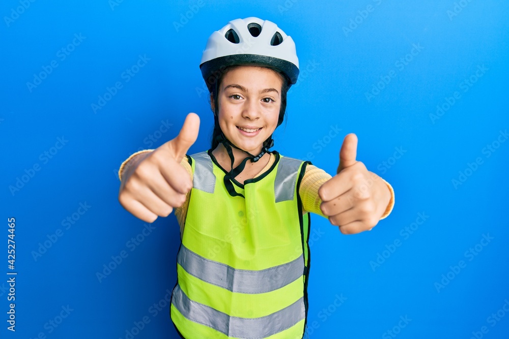 Beautiful brunette little girl wearing bike helmet and reflective vest approving doing positive gesture with hand, thumbs up smiling and happy for success. winner gesture. Beautiful brunette little girl wearing bike helmet and reflective vest approving doing positive gesture with hand, thumbs up smiling and happy for success. winner gesture.