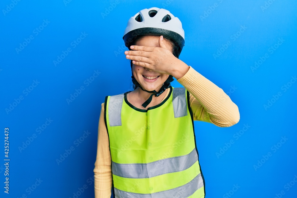 Beautiful brunette little girl wearing bike helmet and reflective vest smiling and laughing with hand on face covering eyes for surprise. blind concept. Beautiful brunette little girl wearing bike helmet and reflective vest smiling and laughing with hand on face covering eyes for surprise. blind concept.