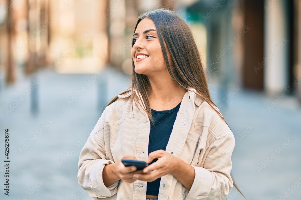 Fototapeta premium Young hispanic girl smiling happy using smartphone at the city.