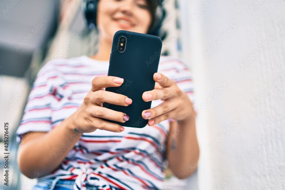 Fototapeta premium Young latin girl smiling happy using smartphone and headphones at the city