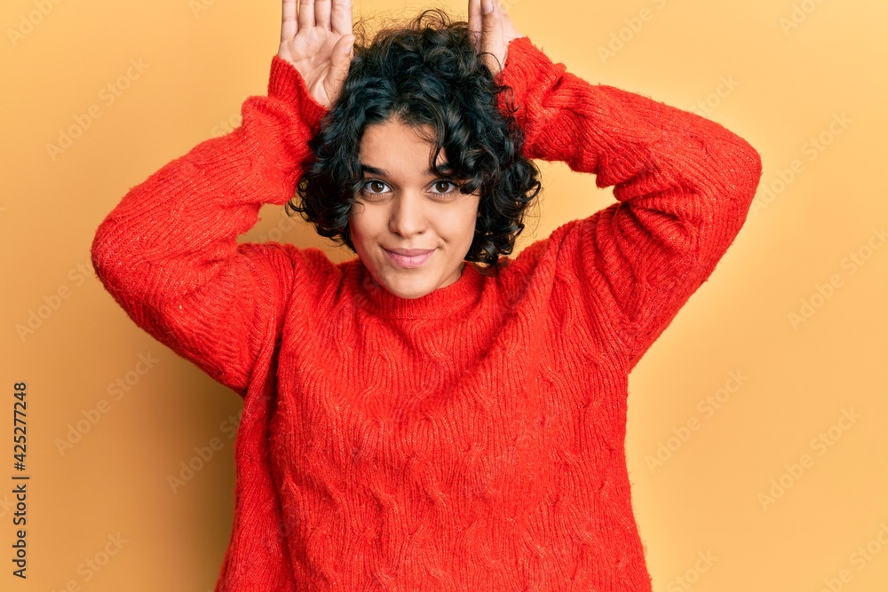 Young hispanic woman with curly hair wearing casual winter sweater doing bunny ears gesture with hands palms looking cynical and skeptical. easter rabbit concept. Young hispanic woman with curly hair wearing casual winter sweater doing bunny ears gesture with hands palms looking cynical and skeptical. easter rabbit concept.