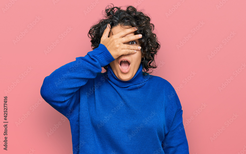 Young hispanic woman with curly hair wearing turtleneck sweater peeking in shock covering face and eyes with hand, looking through fingers with embarrassed expression. Young hispanic woman with curly hair wearing turtleneck sweater peeking in shock covering face and eyes with hand, looking through fingers with embarrassed expression.