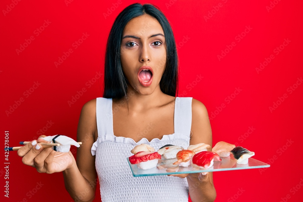 Young brunette woman eating butterfish sushi using chopsticks afraid and shocked with surprise and amazed expression, fear and excited face.
