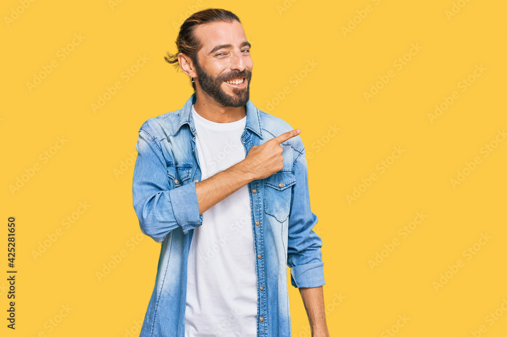 Attractive man with long hair and beard wearing casual denim jacket cheerful with a smile on face pointing with hand and finger up to the side with happy and natural expression