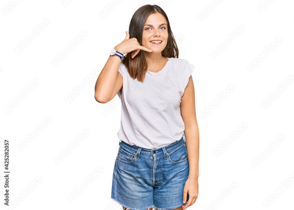 Young caucasian woman wearing casual white tshirt smiling doing phone gesture with hand and fingers like talking on the telephone. communicating concepts. Young caucasian woman wearing casual white tshirt smiling doing phone gesture with hand and fingers like talking on the telephone. communicating concepts.