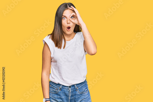 Young caucasian woman wearing casual white tshirt doing ok gesture shocked with surprised face, eye looking through fingers. unbelieving expression.