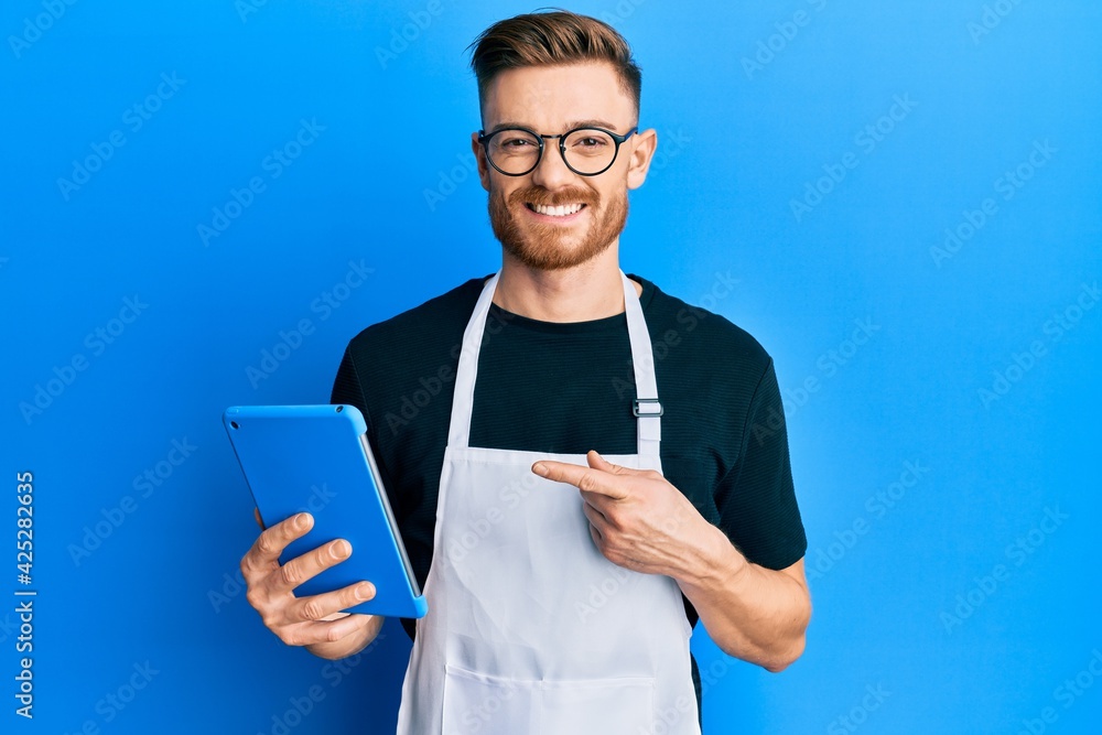 Young redhead man wearing professional apron using touchpad smiling happy pointing with hand and finger Young redhead man wearing professional apron using touchpad smiling happy pointing with hand and finger