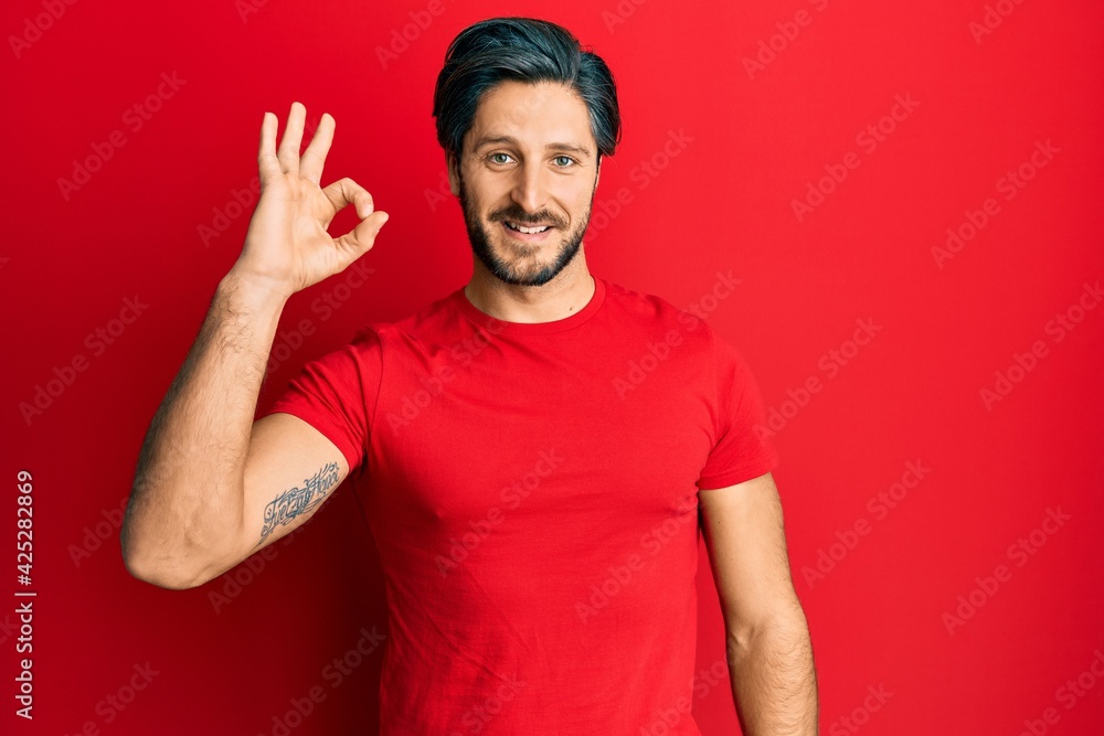 Young hispanic man wearing casual red t shirt smiling positive doing ok sign with hand and fingers. successful expression. Young hispanic man wearing casual red t shirt smiling positive doing ok sign with hand and fingers. successful expression.