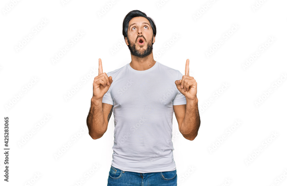 Young hispanic man wearing casual white t shirt amazed and surprised looking up and pointing with fingers and raised arms. Young hispanic man wearing casual white t shirt amazed and surprised looking up and pointing with fingers and raised arms.