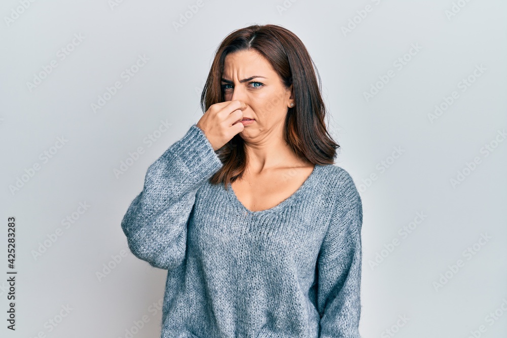Young brunette woman wearing casual winter sweater smelling something stinky and disgusting, intolerable smell, holding breath with fingers on nose. bad smell Young brunette woman wearing casual winter sweater smelling something stinky and disgusting, intolerable smell, holding breath with fingers on nose. bad smell