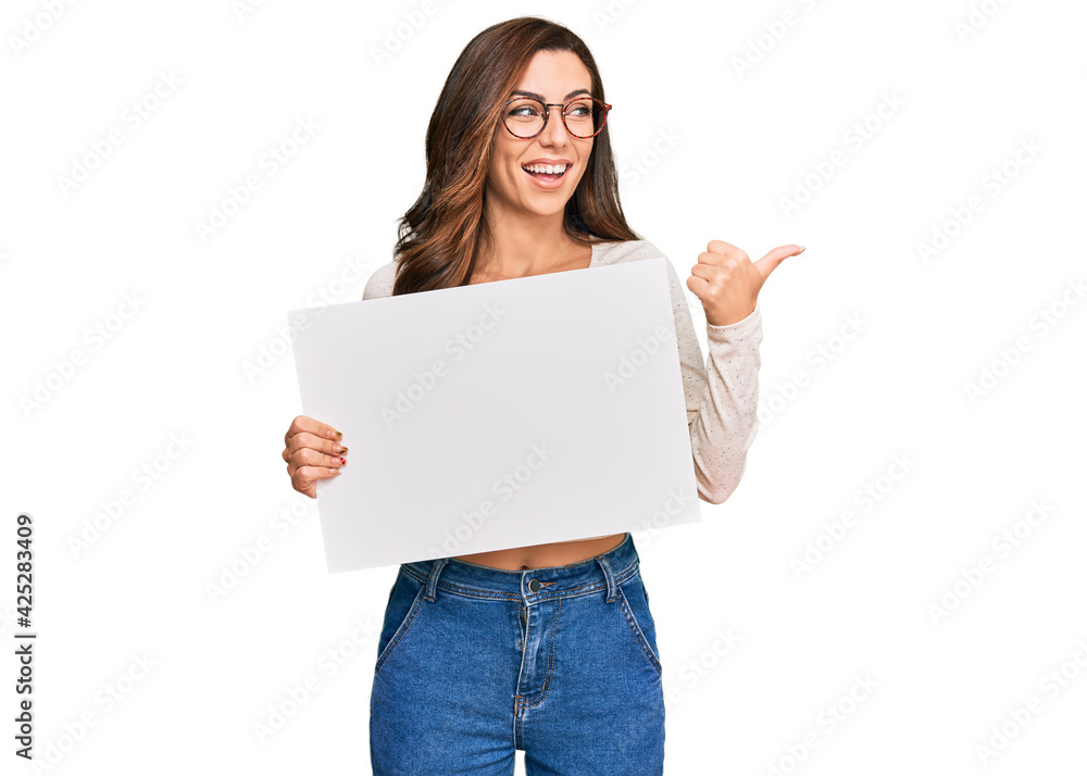 Young brunette woman holding blank empty banner pointing thumb up to the side smiling happy with open mouth Young brunette woman holding blank empty banner pointing thumb up to the side smiling happy with open mouth