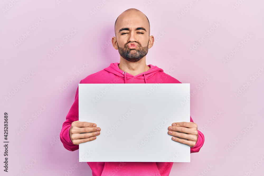 Young bald man holding blank empty banner looking at the camera blowing a kiss being lovely and sexy. love expression. Young bald man holding blank empty banner looking at the camera blowing a kiss being lovely and sexy. love expression.