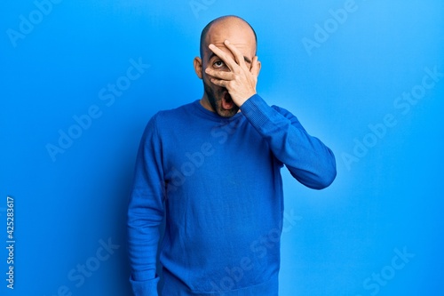 Young hispanic man wearing casual winter sweater peeking in shock covering face and eyes with hand, looking through fingers afraid