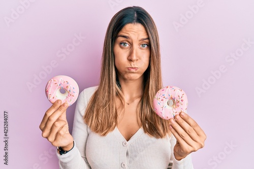 Brunette young woman holding tasty pink doughnuts puffing cheeks with funny face. mouth inflated with air, catching air. photo